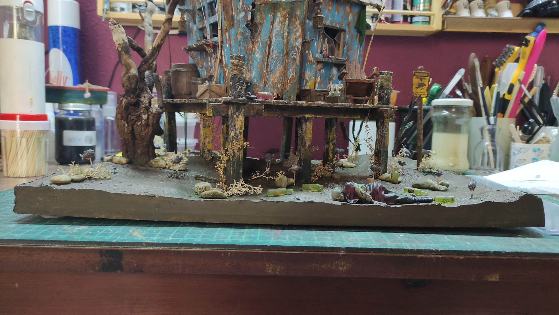 Diorama artístico de una cabaña de madera azul envejecida sobre pilotes en un pantano, con efectos de agua estancada, un bote de remos y detalles de vegetación pantanosa.