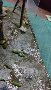 Diorama artístico de una cabaña de madera azul envejecida sobre pilotes en un pantano, con efectos de agua estancada, un bote de remos y detalles de vegetación pantanosa.