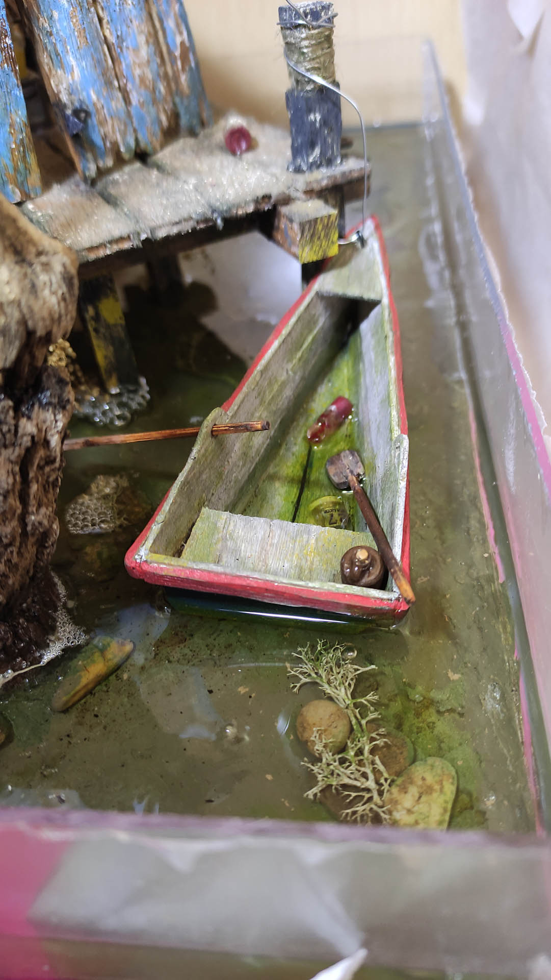Diorama artístico de una cabaña de madera azul envejecida sobre pilotes en un pantano, con efectos de agua estancada, un bote de remos y detalles de vegetación pantanosa.