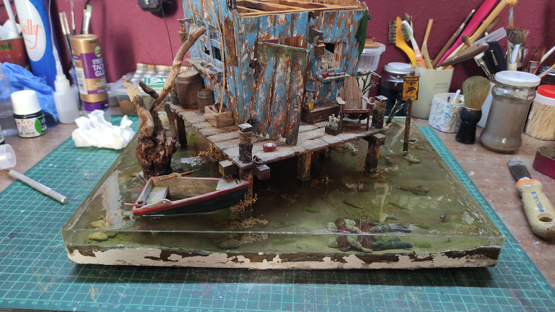 Diorama artístico de una cabaña de madera azul envejecida sobre pilotes en un pantano, con efectos de agua estancada, un bote de remos y detalles de vegetación pantanosa.