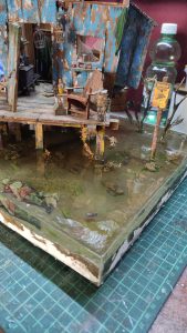 Diorama artístico de una cabaña de madera azul envejecida sobre pilotes en un pantano, con efectos de agua estancada, un bote de remos y detalles de vegetación pantanosa.