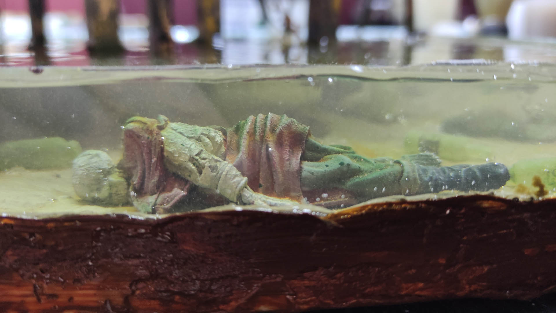 Diorama artístico de una cabaña de madera azul envejecida sobre pilotes en un pantano, con efectos de agua estancada, un bote de remos y detalles de vegetación pantanosa.