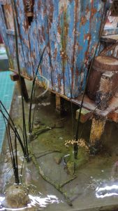 Diorama artístico de una cabaña de madera azul envejecida sobre pilotes en un pantano, con efectos de agua estancada, un bote de remos y detalles de vegetación pantanosa.