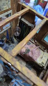 Diorama artístico de una cabaña de madera azul envejecida sobre pilotes en un pantano, con efectos de agua estancada, un bote de remos y detalles de vegetación pantanosa.