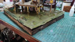 Diorama artístico de una cabaña de madera azul envejecida sobre pilotes en un pantano, con efectos de agua estancada, un bote de remos y detalles de vegetación pantanosa.