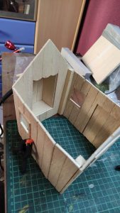 Diorama artístico de una cabaña de madera azul envejecida sobre pilotes en un pantano, con efectos de agua estancada, un bote de remos y detalles de vegetación pantanosa.