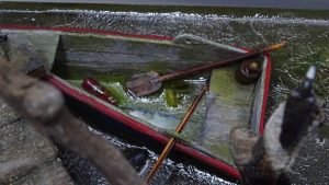 Diorama artístico de una cabaña de madera azul envejecida sobre pilotes en un pantano, con efectos de agua estancada, un bote de remos y detalles de vegetación pantanosa.