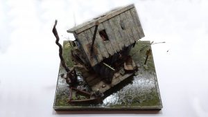 Diorama artístico de una cabaña de madera azul envejecida sobre pilotes en un pantano, con efectos de agua estancada, un bote de remos y detalles de vegetación pantanosa.