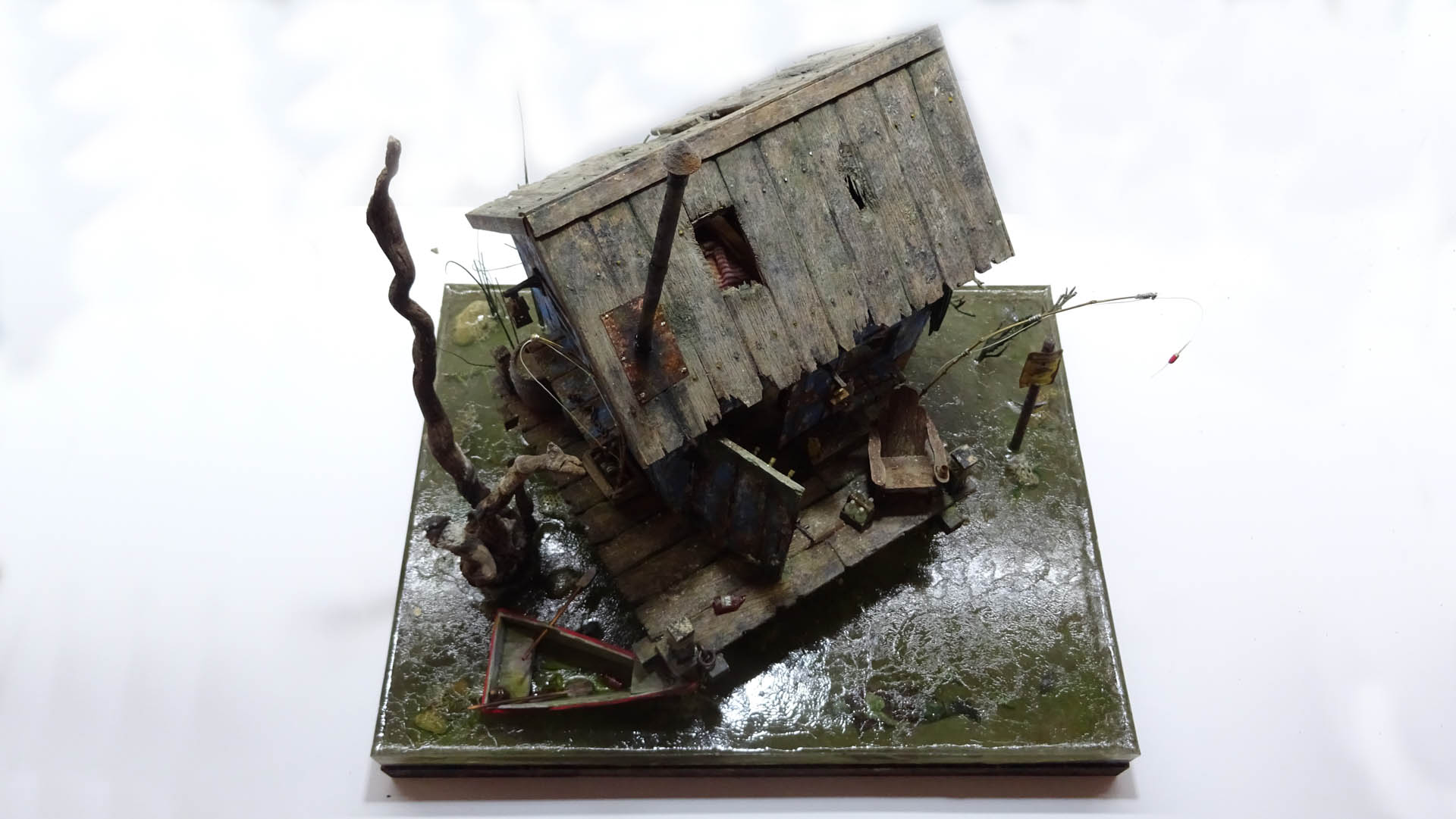 Diorama artístico de una cabaña de madera azul envejecida sobre pilotes en un pantano, con efectos de agua estancada, un bote de remos y detalles de vegetación pantanosa.