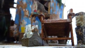 Diorama artístico de una cabaña de madera azul envejecida sobre pilotes en un pantano, con efectos de agua estancada, un bote de remos y detalles de vegetación pantanosa.