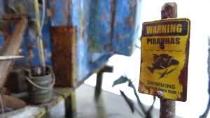 Diorama artístico de una cabaña de madera azul envejecida sobre pilotes en un pantano, con efectos de agua estancada, un bote de remos y detalles de vegetación pantanosa.