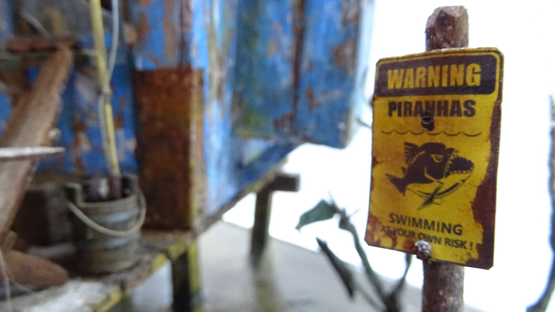 Diorama artístico de una cabaña de madera azul envejecida sobre pilotes en un pantano, con efectos de agua estancada, un bote de remos y detalles de vegetación pantanosa.