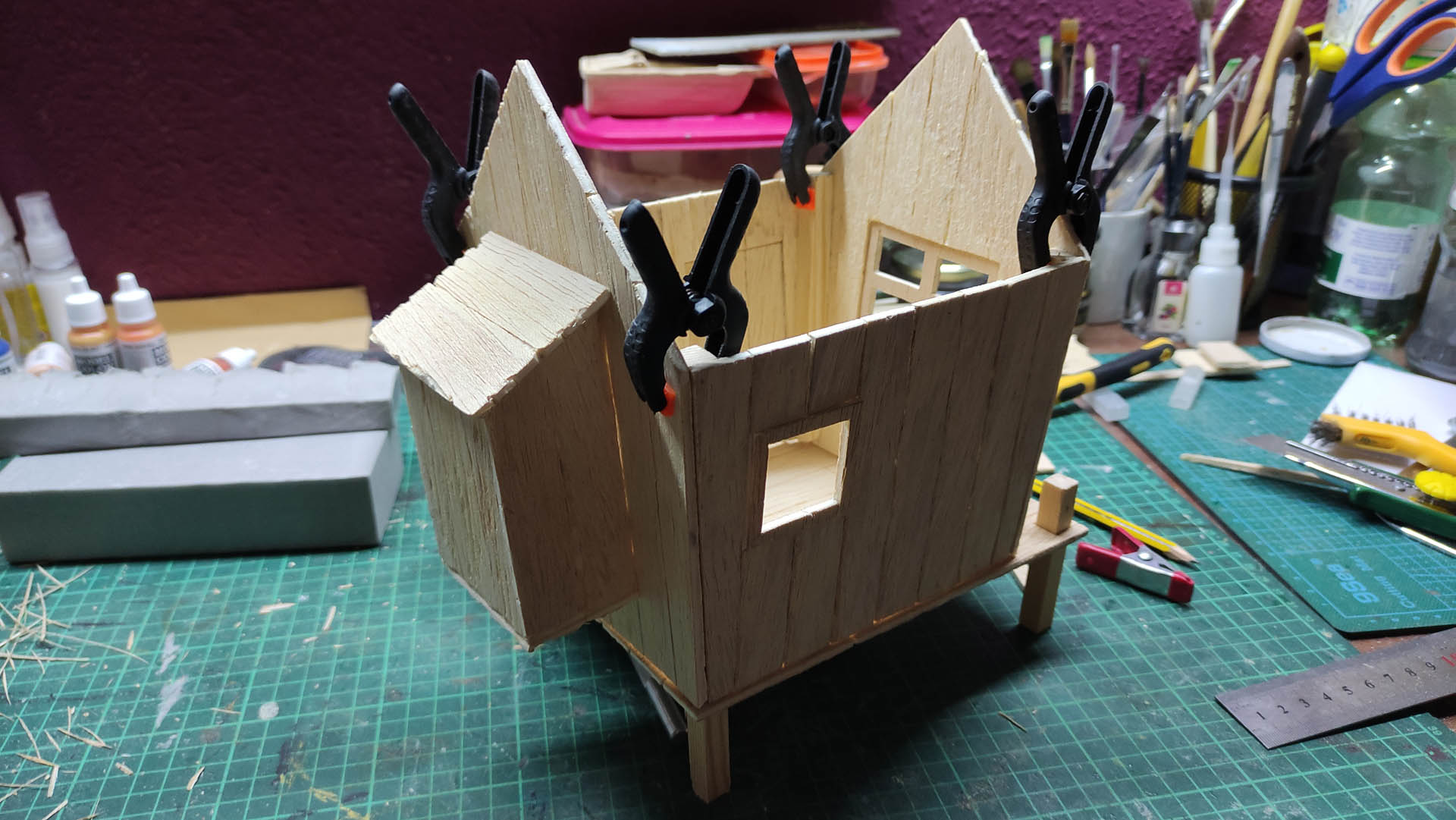 Diorama artístico de una cabaña de madera azul envejecida sobre pilotes en un pantano, con efectos de agua estancada, un bote de remos y detalles de vegetación pantanosa.