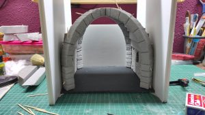 Diorama artesanal detallado de un duende y un pozo estructurado en tres niveles. El nivel superior recrea un jardín exterior exuberante utilizando vegetación natural y preservada (musgos y enredaderas), sobre estancias subterráneas de piedra y madera