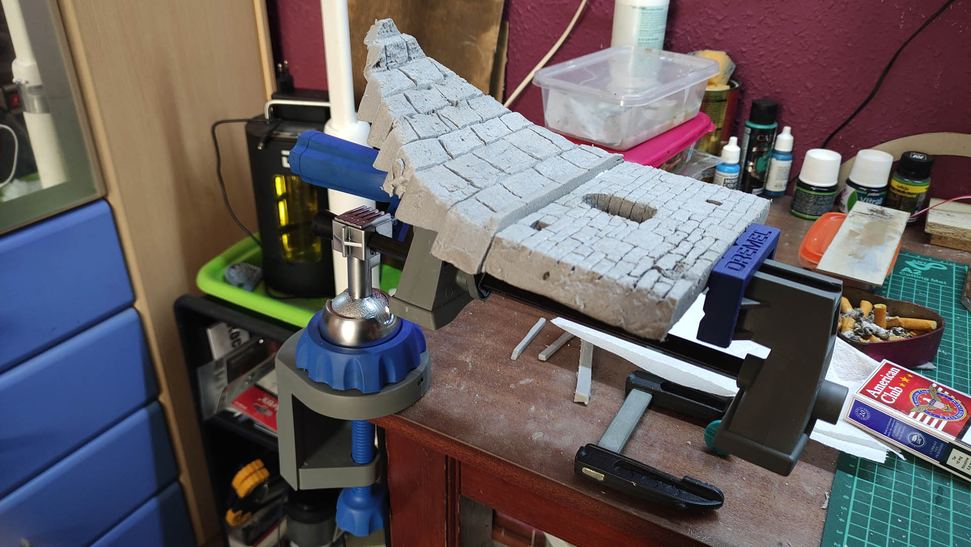 Diorama artesanal detallado de un duende y un pozo estructurado en tres niveles. El nivel superior recrea un jardín exterior exuberante utilizando vegetación natural y preservada (musgos y enredaderas), sobre estancias subterráneas de piedra y madera