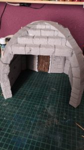 Diorama artesanal detallado de un duende y un pozo estructurado en tres niveles. El nivel superior recrea un jardín exterior exuberante utilizando vegetación natural y preservada (musgos y enredaderas), sobre estancias subterráneas de piedra y madera