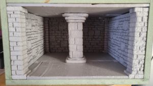 Diorama artesanal detallado de un duende y un pozo estructurado en tres niveles. El nivel superior recrea un jardín exterior exuberante utilizando vegetación natural y preservada (musgos y enredaderas), sobre estancias subterráneas de piedra y madera