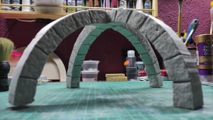 Diorama artesanal detallado de un duende y un pozo estructurado en tres niveles. El nivel superior recrea un jardín exterior exuberante utilizando vegetación natural y preservada (musgos y enredaderas), sobre estancias subterráneas de piedra y madera