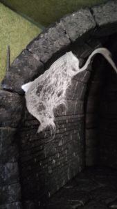 Diorama artesanal detallado de un duende y un pozo estructurado en tres niveles. El nivel superior recrea un jardín exterior exuberante utilizando vegetación natural y preservada (musgos y enredaderas), sobre estancias subterráneas de piedra y madera