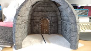 Diorama artesanal detallado de un duende y un pozo estructurado en tres niveles. El nivel superior recrea un jardín exterior exuberante utilizando vegetación natural y preservada (musgos y enredaderas), sobre estancias subterráneas de piedra y madera