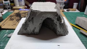 Diorama artesanal detallado de un duende y un pozo estructurado en tres niveles. El nivel superior recrea un jardín exterior exuberante utilizando vegetación natural y preservada (musgos y enredaderas), sobre estancias subterráneas de piedra y madera