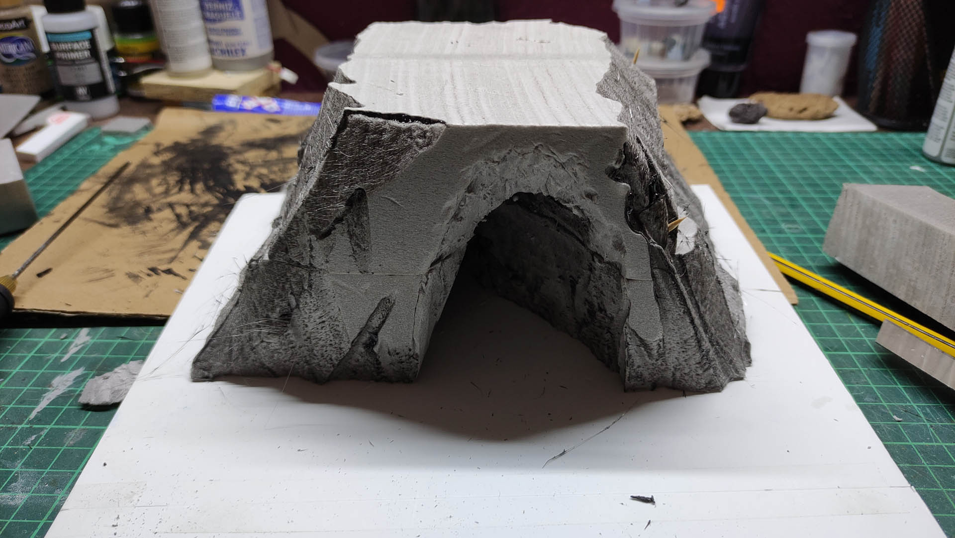 Diorama artesanal detallado de un duende y un pozo estructurado en tres niveles. El nivel superior recrea un jardín exterior exuberante utilizando vegetación natural y preservada (musgos y enredaderas), sobre estancias subterráneas de piedra y madera