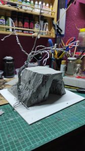 Diorama artesanal detallado de un duende y un pozo estructurado en tres niveles. El nivel superior recrea un jardín exterior exuberante utilizando vegetación natural y preservada (musgos y enredaderas), sobre estancias subterráneas de piedra y madera