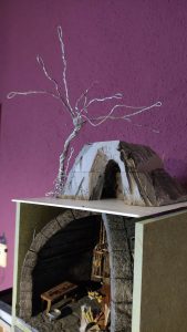 Diorama artesanal detallado de un duende y un pozo estructurado en tres niveles. El nivel superior recrea un jardín exterior exuberante utilizando vegetación natural y preservada (musgos y enredaderas), sobre estancias subterráneas de piedra y madera