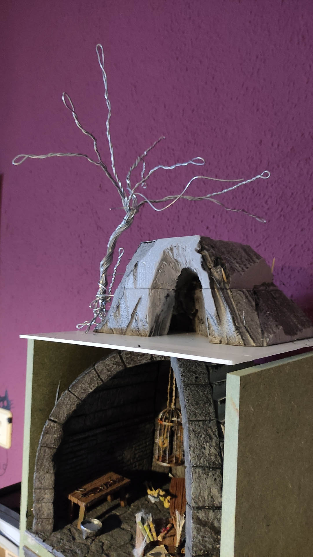 Diorama artesanal detallado de un duende y un pozo estructurado en tres niveles. El nivel superior recrea un jardín exterior exuberante utilizando vegetación natural y preservada (musgos y enredaderas), sobre estancias subterráneas de piedra y madera