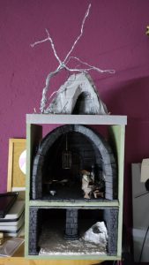 Diorama artesanal detallado de un duende y un pozo estructurado en tres niveles. El nivel superior recrea un jardín exterior exuberante utilizando vegetación natural y preservada (musgos y enredaderas), sobre estancias subterráneas de piedra y madera