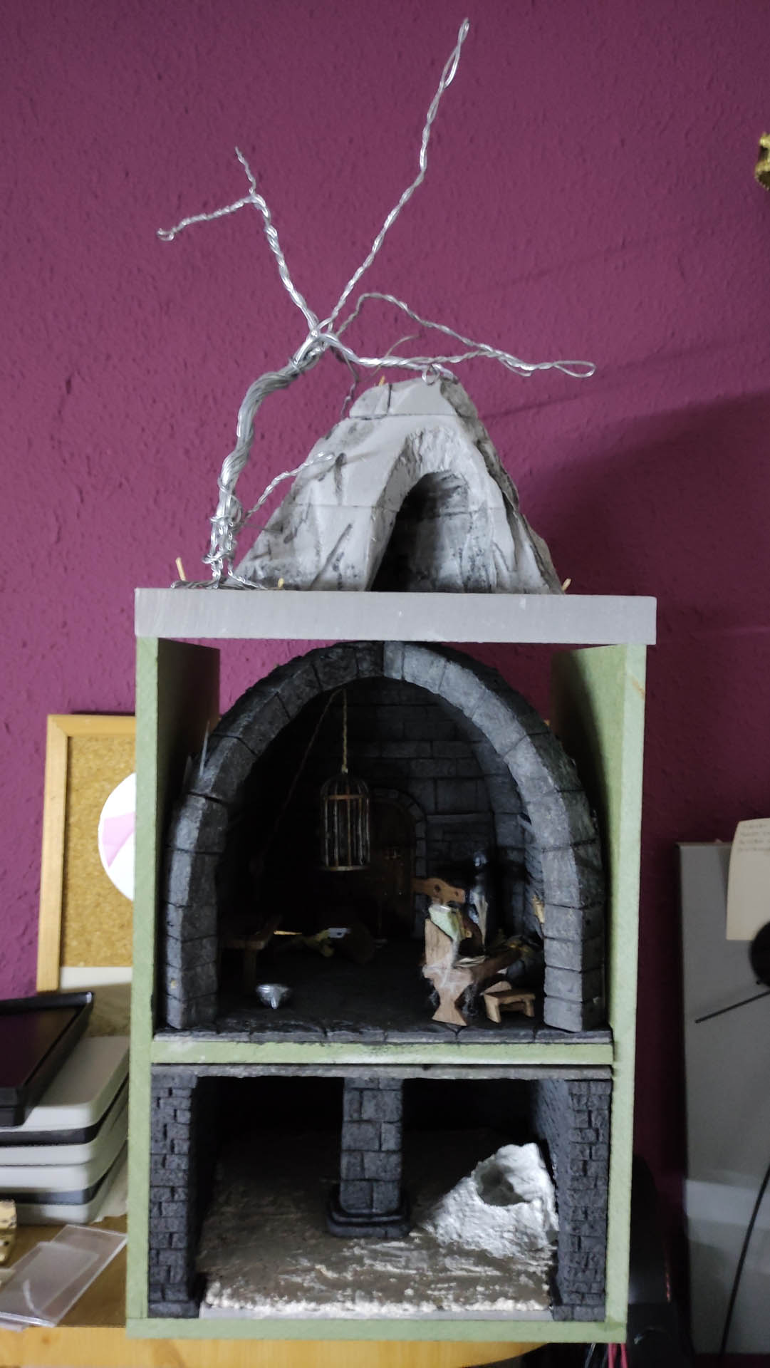 Diorama artesanal detallado de un duende y un pozo estructurado en tres niveles. El nivel superior recrea un jardín exterior exuberante utilizando vegetación natural y preservada (musgos y enredaderas), sobre estancias subterráneas de piedra y madera