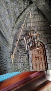 Diorama artesanal detallado de un duende y un pozo estructurado en tres niveles. El nivel superior recrea un jardín exterior exuberante utilizando vegetación natural y preservada (musgos y enredaderas), sobre estancias subterráneas de piedra y madera