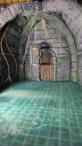 Diorama artesanal detallado de un duende y un pozo estructurado en tres niveles. El nivel superior recrea un jardín exterior exuberante utilizando vegetación natural y preservada (musgos y enredaderas), sobre estancias subterráneas de piedra y madera