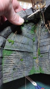 Diorama artesanal detallado de un duende y un pozo estructurado en tres niveles. El nivel superior recrea un jardín exterior exuberante utilizando vegetación natural y preservada (musgos y enredaderas), sobre estancias subterráneas de piedra y madera