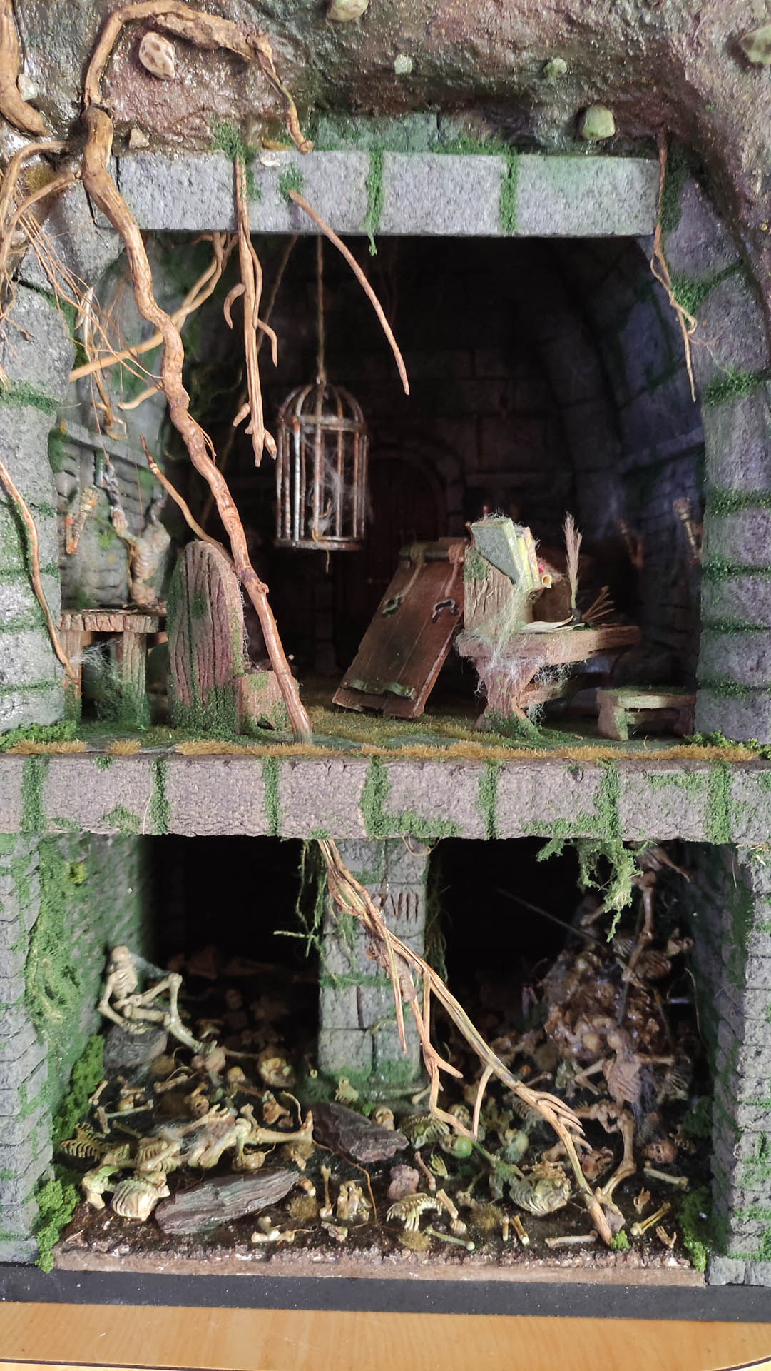 Diorama artesanal detallado de un duende y un pozo estructurado en tres niveles. El nivel superior recrea un jardín exterior exuberante utilizando vegetación natural y preservada (musgos y enredaderas), sobre estancias subterráneas de piedra y madera