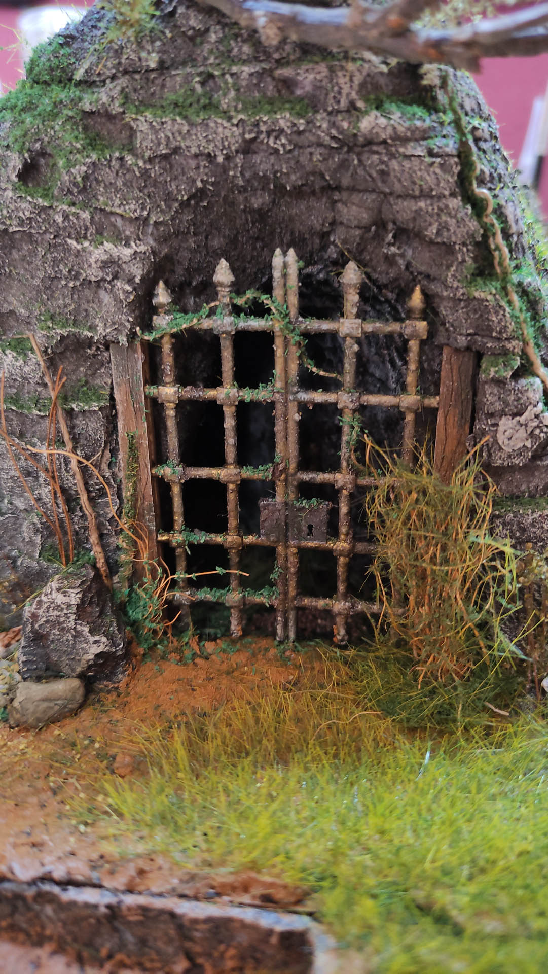 Diorama artesanal detallado de un duende y un pozo estructurado en tres niveles. El nivel superior recrea un jardín exterior exuberante utilizando vegetación natural y preservada (musgos y enredaderas), sobre estancias subterráneas de piedra y madera