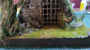 Diorama artesanal detallado de un duende y un pozo estructurado en tres niveles. El nivel superior recrea un jardín exterior exuberante utilizando vegetación natural y preservada (musgos y enredaderas), sobre estancias subterráneas de piedra y madera