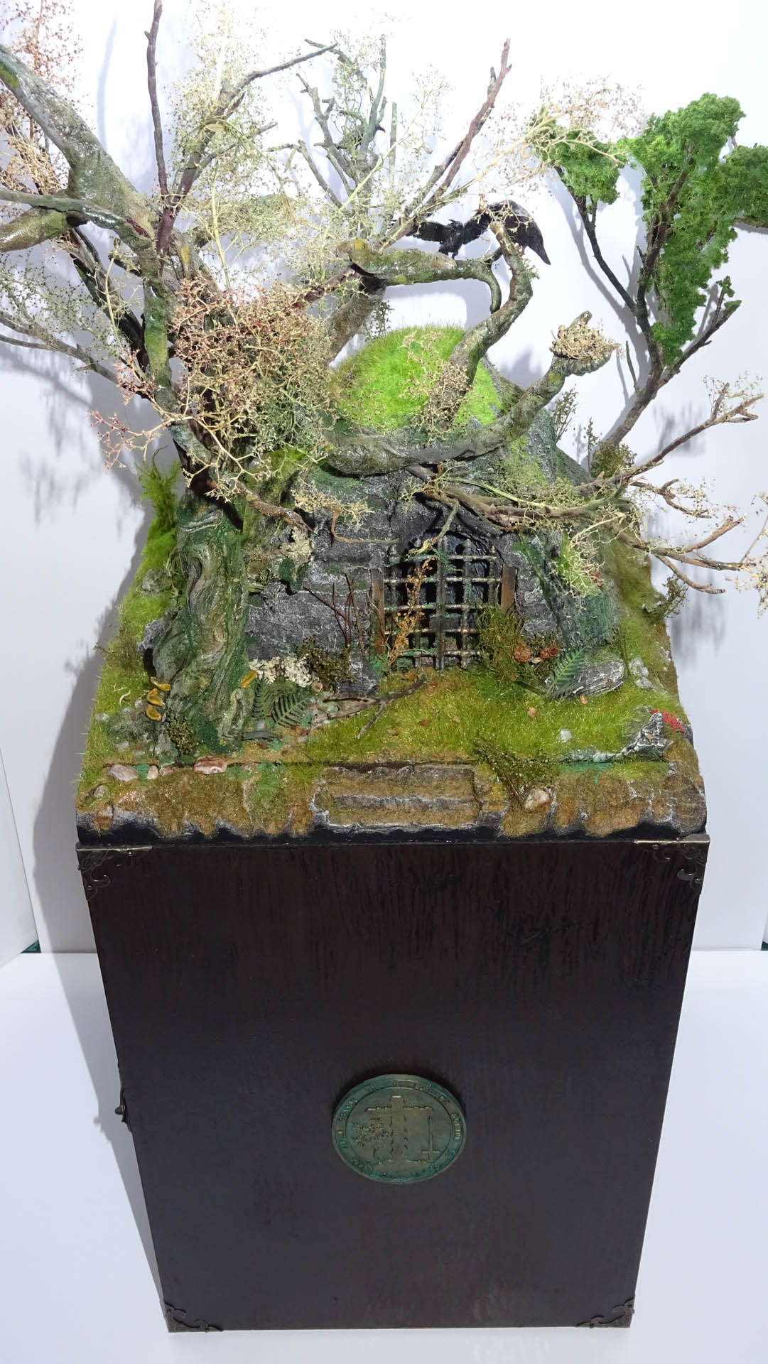 Diorama artesanal detallado de un duende y un pozo estructurado en tres niveles. El nivel superior recrea un jardín exterior exuberante utilizando vegetación natural y preservada (musgos y enredaderas), sobre estancias subterráneas de piedra y madera