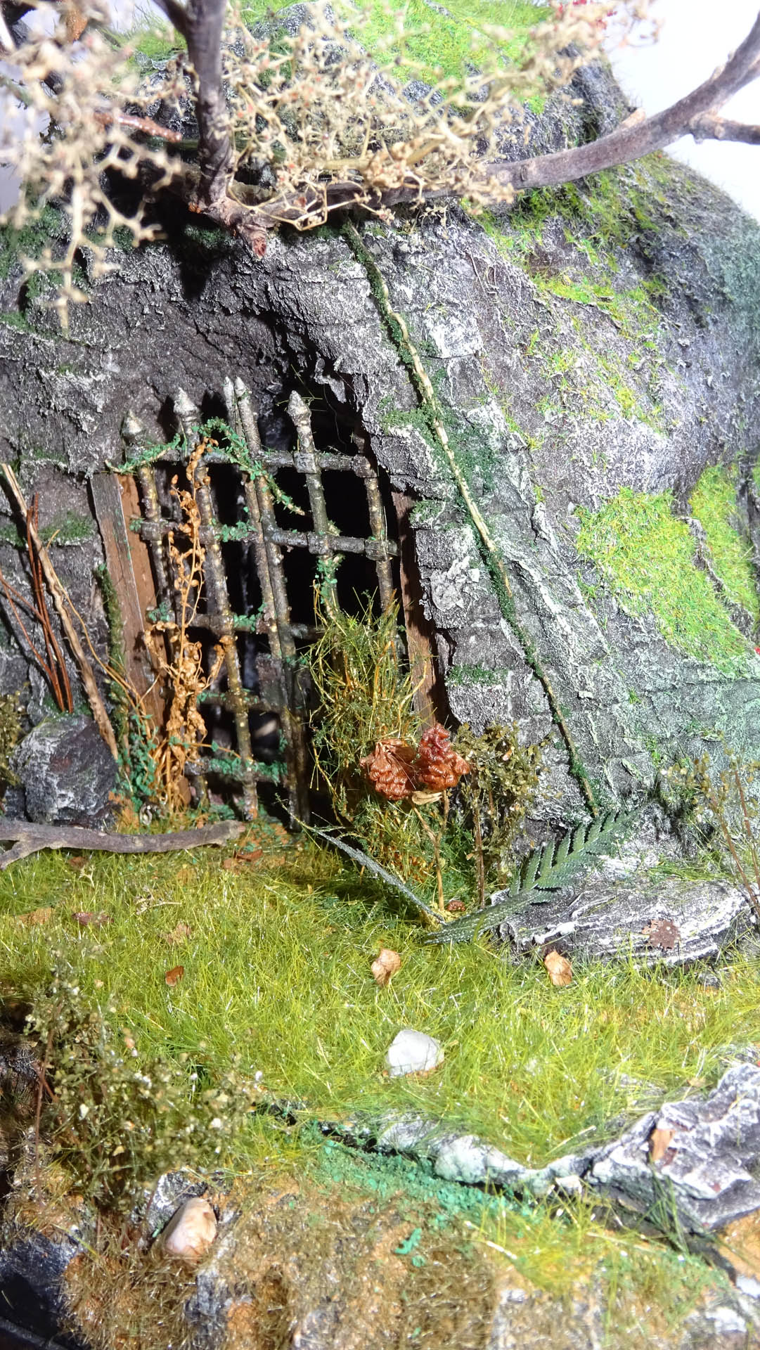 Diorama artesanal detallado de un duende y un pozo estructurado en tres niveles. El nivel superior recrea un jardín exterior exuberante utilizando vegetación natural y preservada (musgos y enredaderas), sobre estancias subterráneas de piedra y madera