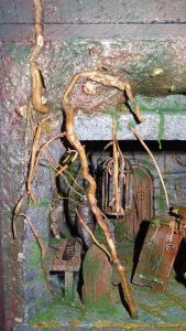 Diorama artesanal detallado de un duende y un pozo estructurado en tres niveles. El nivel superior recrea un jardín exterior exuberante utilizando vegetación natural y preservada (musgos y enredaderas), sobre estancias subterráneas de piedra y madera