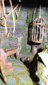 Diorama artesanal detallado de un duende y un pozo estructurado en tres niveles. El nivel superior recrea un jardín exterior exuberante utilizando vegetación natural y preservada (musgos y enredaderas), sobre estancias subterráneas de piedra y madera