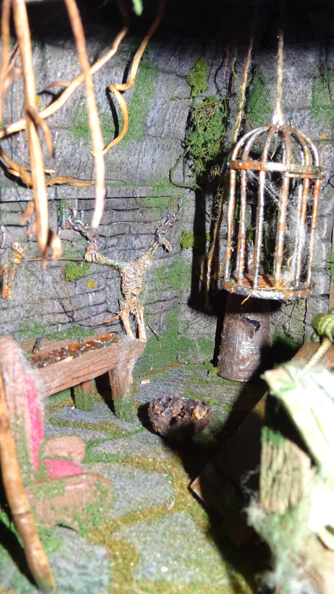 Diorama artesanal detallado de un duende y un pozo estructurado en tres niveles. El nivel superior recrea un jardín exterior exuberante utilizando vegetación natural y preservada (musgos y enredaderas), sobre estancias subterráneas de piedra y madera