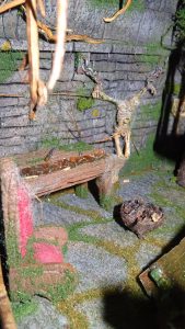 Diorama artesanal detallado de un duende y un pozo estructurado en tres niveles. El nivel superior recrea un jardín exterior exuberante utilizando vegetación natural y preservada (musgos y enredaderas), sobre estancias subterráneas de piedra y madera