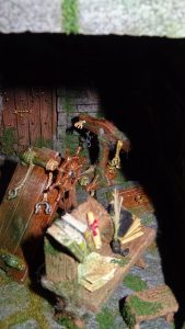 Diorama artesanal detallado de un duende y un pozo estructurado en tres niveles. El nivel superior recrea un jardín exterior exuberante utilizando vegetación natural y preservada (musgos y enredaderas), sobre estancias subterráneas de piedra y madera