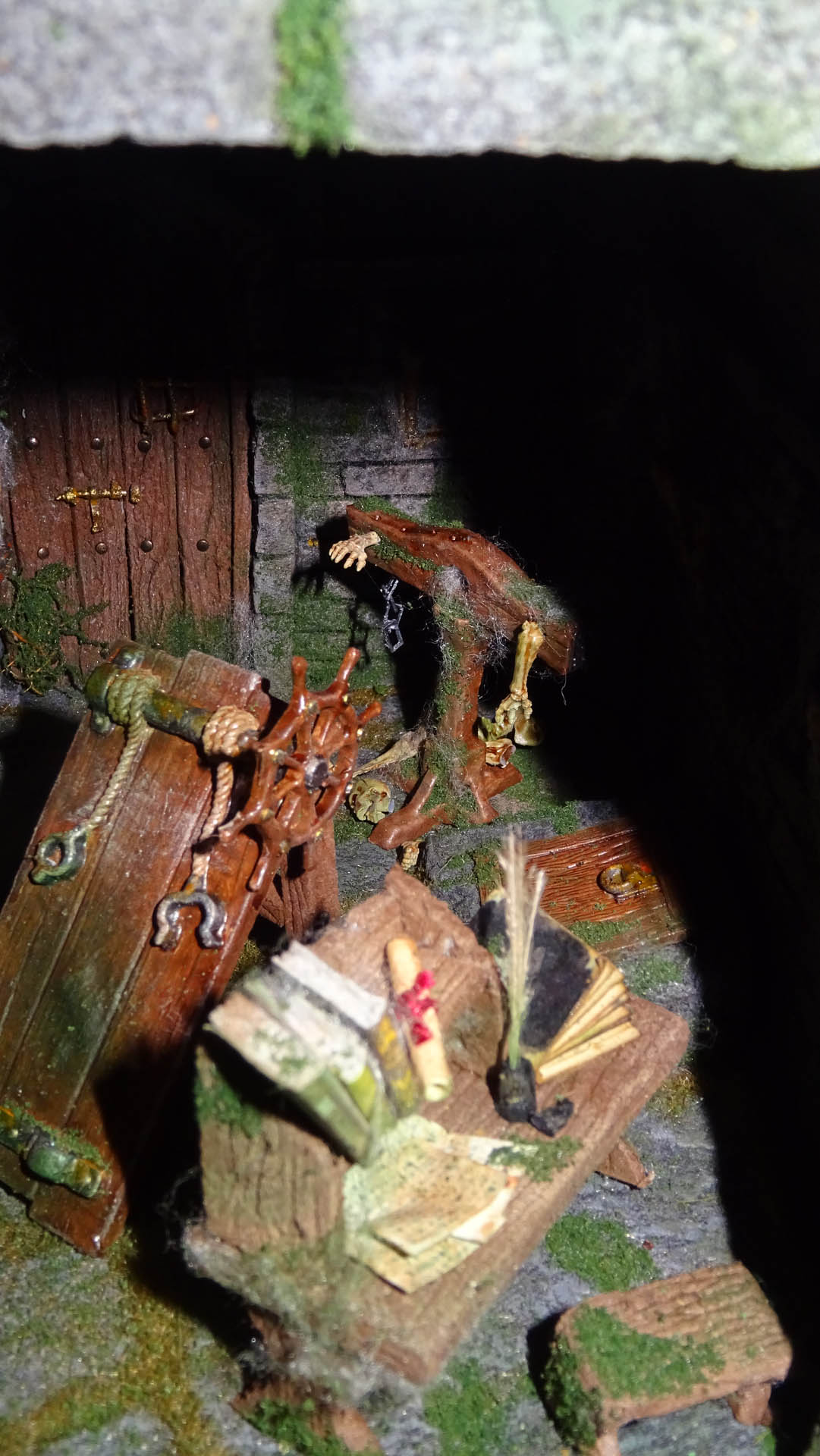 Diorama artesanal detallado de un duende y un pozo estructurado en tres niveles. El nivel superior recrea un jardín exterior exuberante utilizando vegetación natural y preservada (musgos y enredaderas), sobre estancias subterráneas de piedra y madera