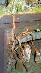 Diorama artesanal detallado de un duende y un pozo estructurado en tres niveles. El nivel superior recrea un jardín exterior exuberante utilizando vegetación natural y preservada (musgos y enredaderas), sobre estancias subterráneas de piedra y madera