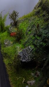 Diorama artesanal detallado de un duende y un pozo estructurado en tres niveles. El nivel superior recrea un jardín exterior exuberante utilizando vegetación natural y preservada (musgos y enredaderas), sobre estancias subterráneas de piedra y madera