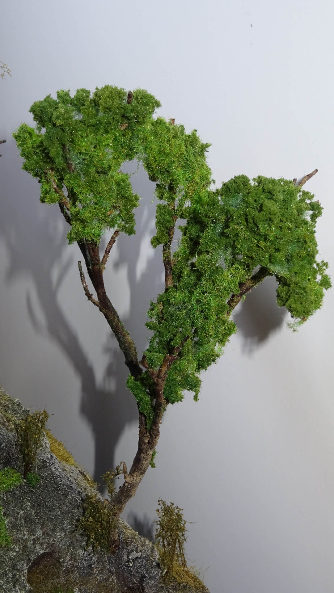 Diorama artesanal detallado de un duende y un pozo estructurado en tres niveles. El nivel superior recrea un jardín exterior exuberante utilizando vegetación natural y preservada (musgos y enredaderas), sobre estancias subterráneas de piedra y madera