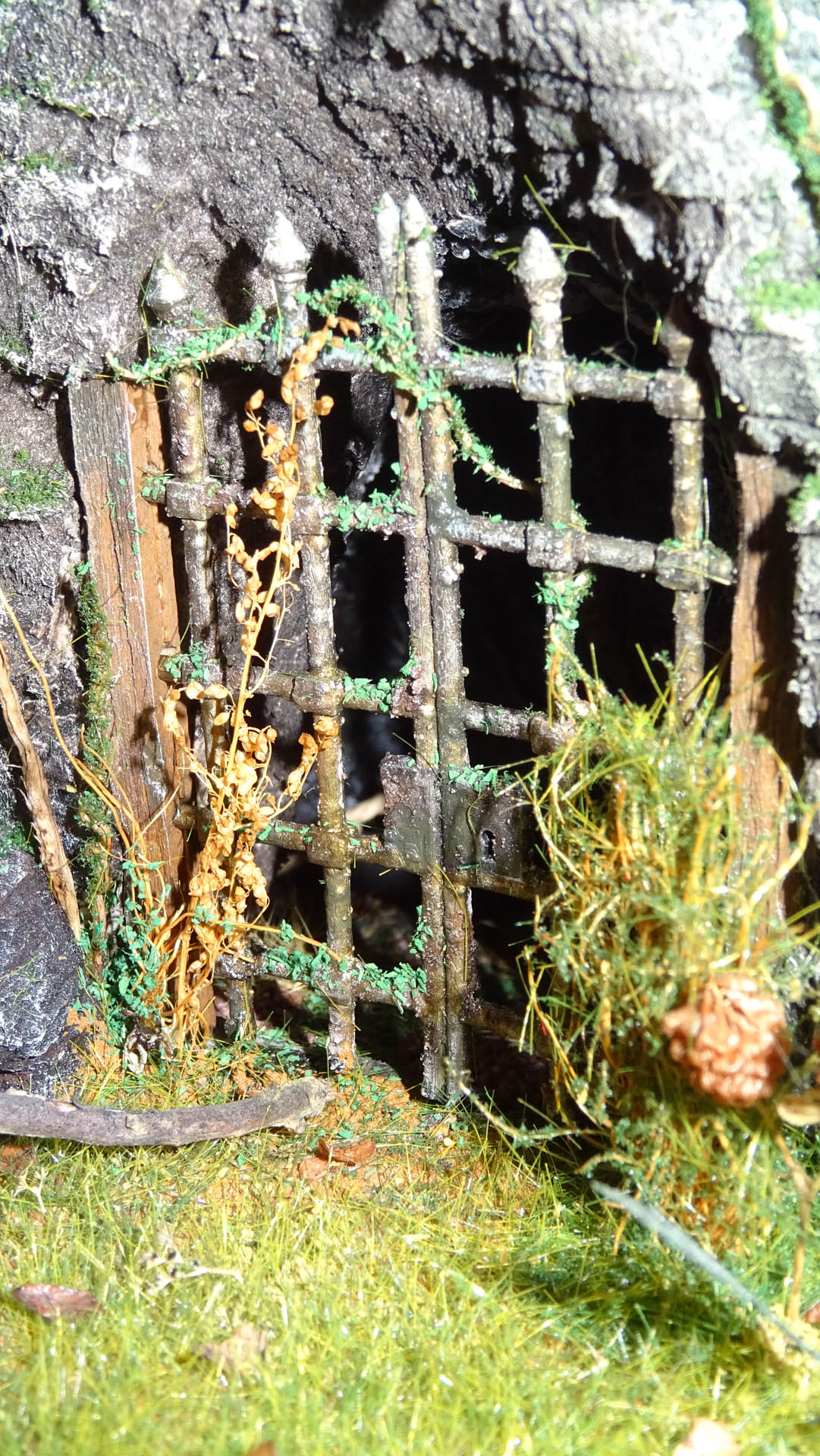 Diorama artesanal detallado de un duende y un pozo estructurado en tres niveles. El nivel superior recrea un jardín exterior exuberante utilizando vegetación natural y preservada (musgos y enredaderas), sobre estancias subterráneas de piedra y madera