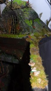 Diorama artesanal detallado de un duende y un pozo estructurado en tres niveles. El nivel superior recrea un jardín exterior exuberante utilizando vegetación natural y preservada (musgos y enredaderas), sobre estancias subterráneas de piedra y madera